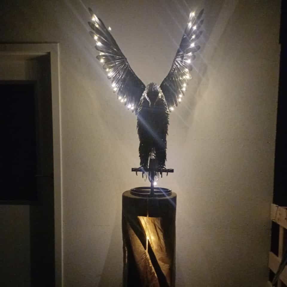 Skulptur eines Vogels mit leuchtenden Flügeln auf einem Holzsockel.